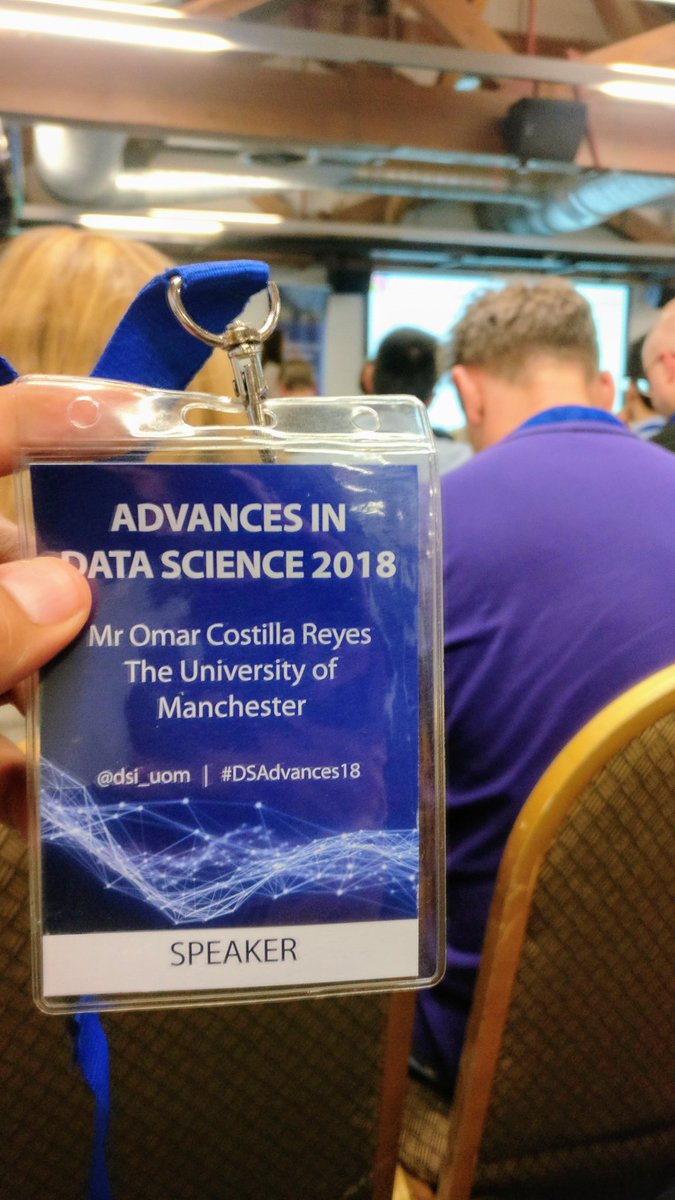 Hand holding a blue Advances in Data Science 2018 speaker badge that reads Mr Omar Costilla Reyes, The University of Manchester, with an audience seated in the room behind it.
