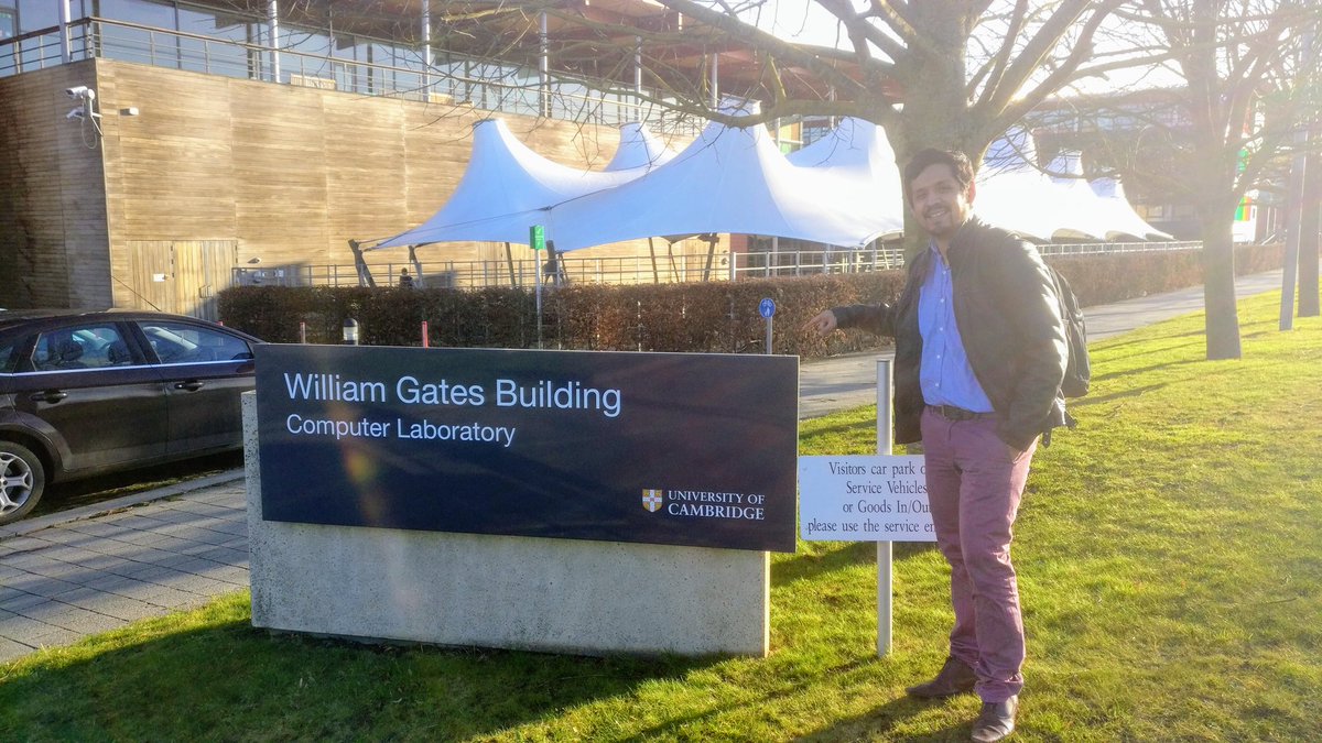 A man stands beside the William Gates Building Computer Laboratory sign of the University of Cambridge, with white tent canopies and a building behind.