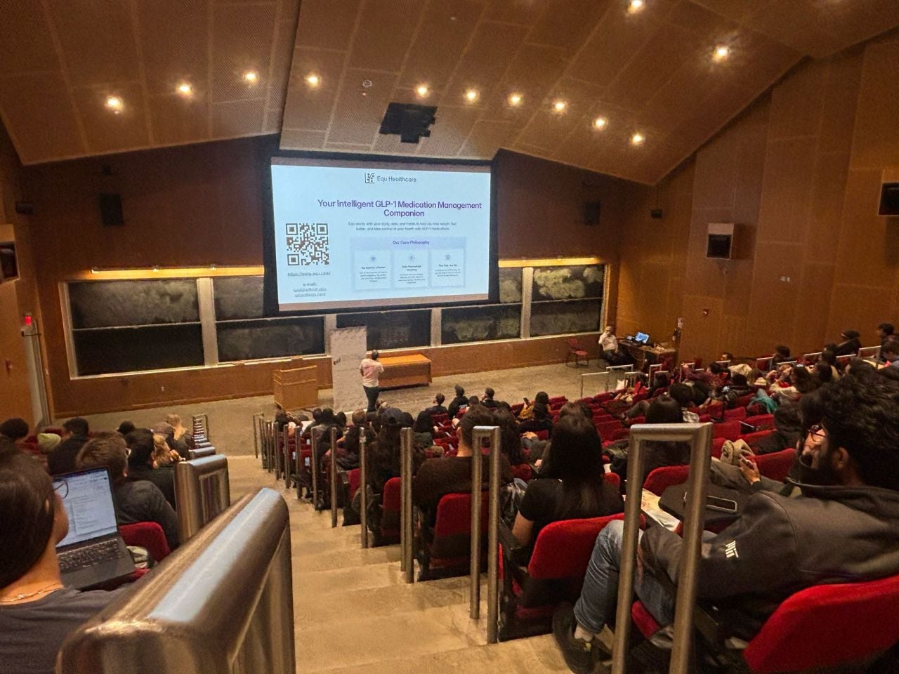 A tiered lecture hall with an audience facing a screen that shows an Equ Healthcare slide titled “Your Intelligent GLP-1 Medication Management Companion,” with a QR code.