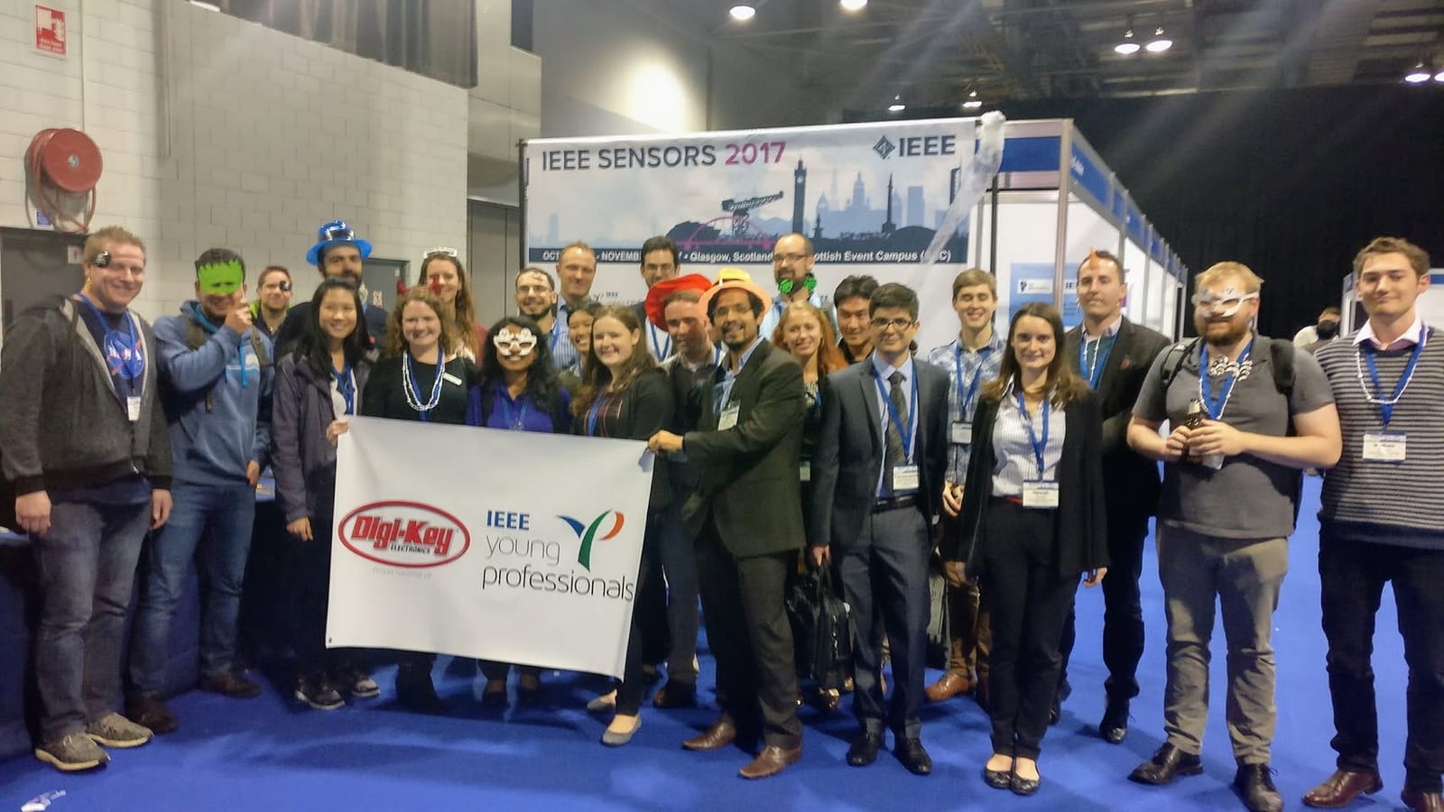 About two dozen conference attendees, a few in masks or hats, hold a Digi-Key and IEEE Young Professionals banner in front of an IEEE SENSORS 2017 Glasgow sign.