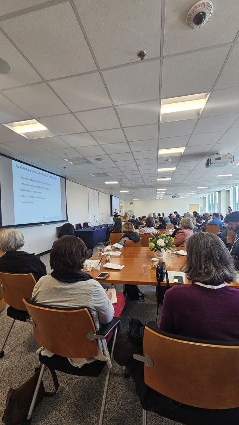 Symposium attendees seated at long tables facing a projected slide on the spatial dimensions of collective well-being.