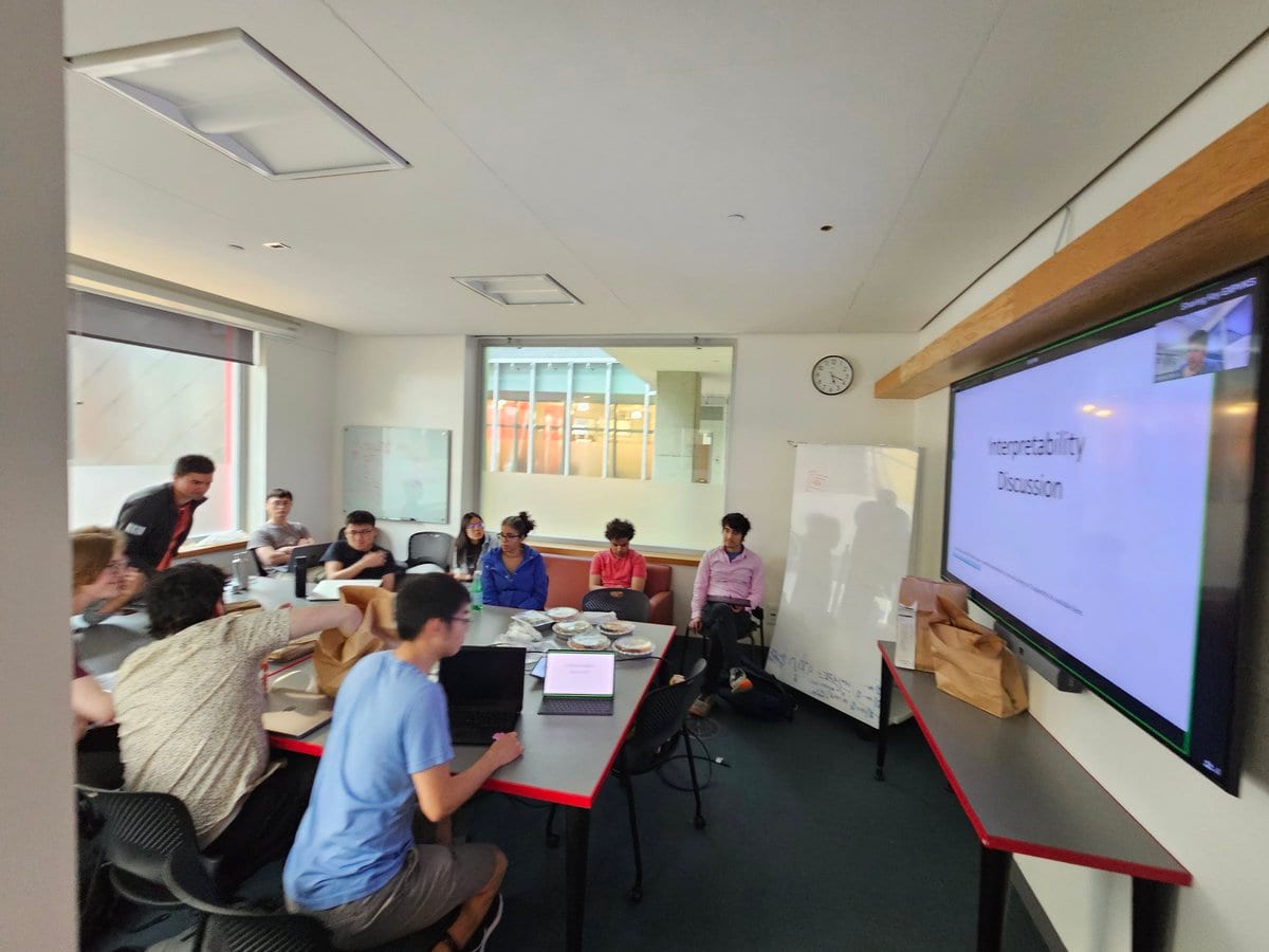 Participants seated around a table and on a couch in a meeting room face a wall screen that reads “Interpretability Discussion”, with a video-call participant in the corner.