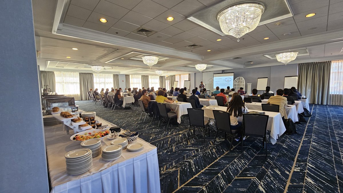 Participants seated at round tables in a ballroom-style conference room facing a presentation screen, with a food table in the foreground.