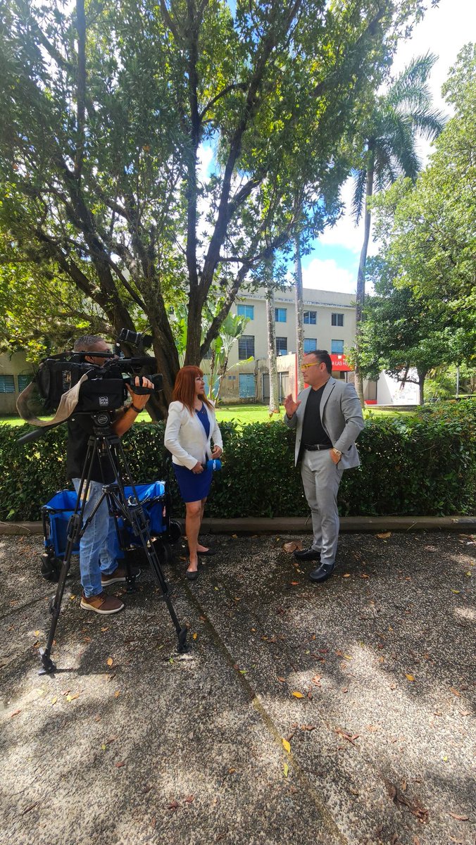 A television camera crew filming an interview with adults in a campus courtyard.