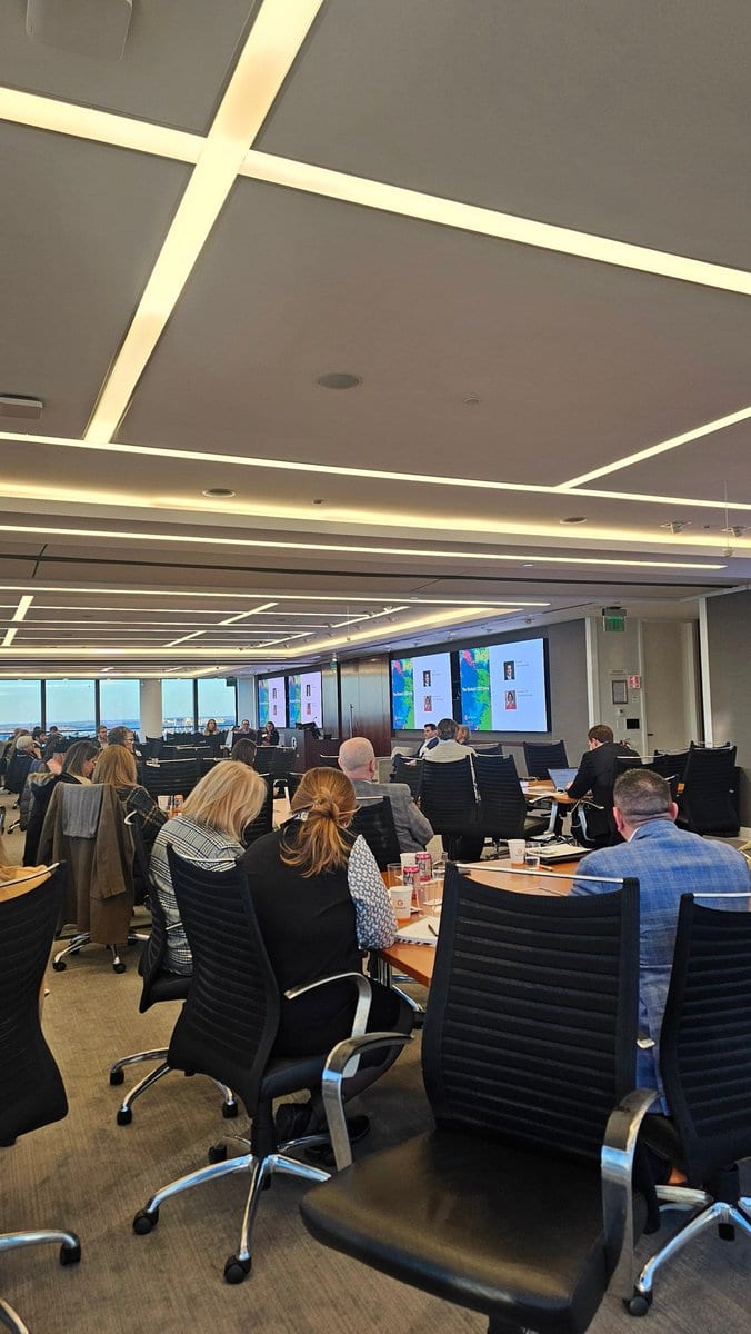 Attendees sit at tables in a conference room with large windows, facing wall screens that show speaker slides during a symposium session.