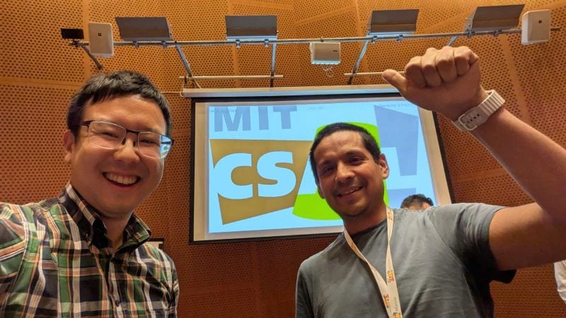 Two smiling people take a close selfie in a lecture hall in front of a projection screen showing the MIT CSAIL logo.