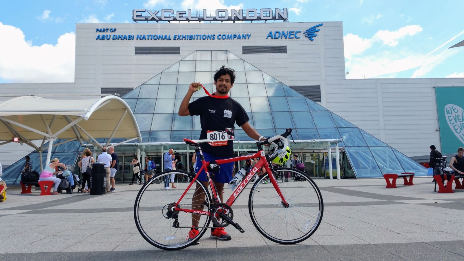 Holding up my London Triathlon medal beside my red road bike, wearing race number 8016, outside ExCeL London.