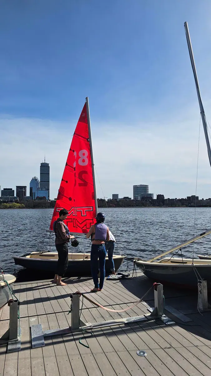 A red MIT sailing dinghy at the dock with Boston across the river.